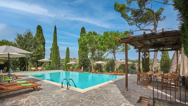 A swimming pool with lounge chairs and umbrellas, surrounded by stone flooring and tall trees. - Apartment with terrace and enchanting view (Torrita di Siena)