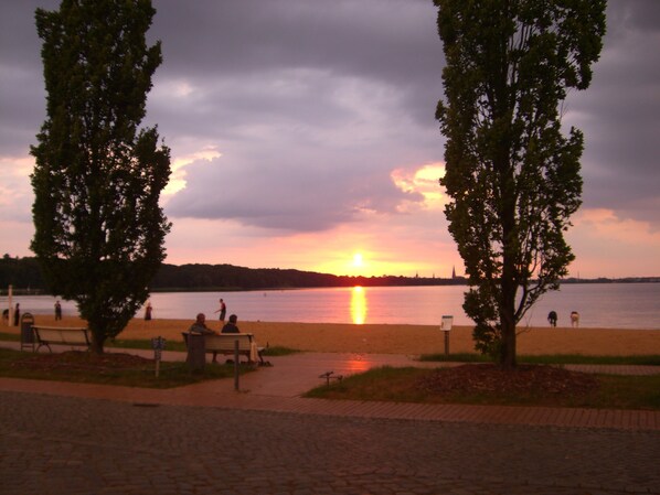 A sunset view over a lake with a park area, benches, and trees. - Apartment beach time, lovingly furnished, quiet location on Lake Schwerin (Schwerin - Zippendorf)