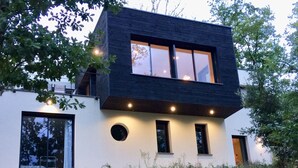 Une maison moderne à deux étages, dotée de grandes fenêtres, d’un toit plat et d’une façade blanche.