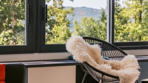 Une pièce moderne avec une grande fenêtre, une chaise noire dotée d’un coussin blanc moelleux et un oreiller rouge sur un canapé sombre.