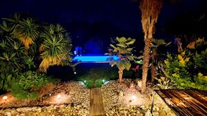 Ein Garten bei Nacht mit einem Weg, der zu einem Poolbereich führt, umgeben von Palmen und verschiedenen Pflanzen.