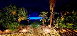 A nighttime garden with a pathway leading to a pool area, surrounded by palm trees and various plants. - 700 meters from the ocean, House with volleyball, pétanque, badminton, ping-pong, and swings (Santa-Lucia-Di-Moriani)