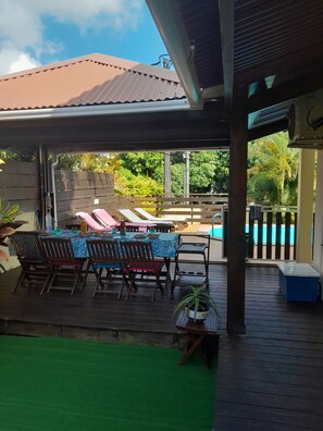 Un espacio exterior techado con piscina, terraza de madera y sillas de descanso.