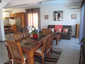 Un comedor con una mesa y sillas de madera, una cocina al fondo y una sala de estar con un sofá y un cuadro en la pared.