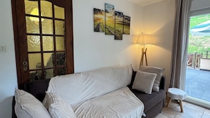 A modern living room with a dark grey sofa, a wooden door with glass panels, a floor lamp, and a view of the outdoors through a large window.