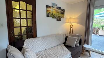 A modern living room with a dark grey sofa, a wooden door with glass panels, a floor lamp, and a view of the outdoors through a large window.