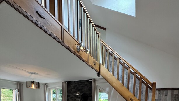 A spacious dining area with a high wooden staircase, a stone fireplace, and large windows offering a view of the outdoors.