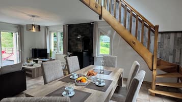 A spacious dining area with a high wooden staircase, a stone fireplace, and large windows offering a view of the outdoors.
