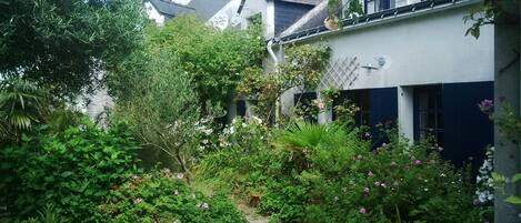 Un jardin luxuriant, avec une végétation dense et des plantes en fleurs, se trouve devant un bâtiment à la façade blanche et aux volets sombres.
