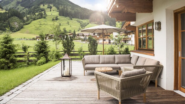 Una terraza de madera con un sofá y sillas de mimbre, con vistas a un valle verde y montañas.
