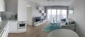 A modern living room with a flat-screen TV, a sofa, a coffee table, and a kitchen area with a stove and a sink.