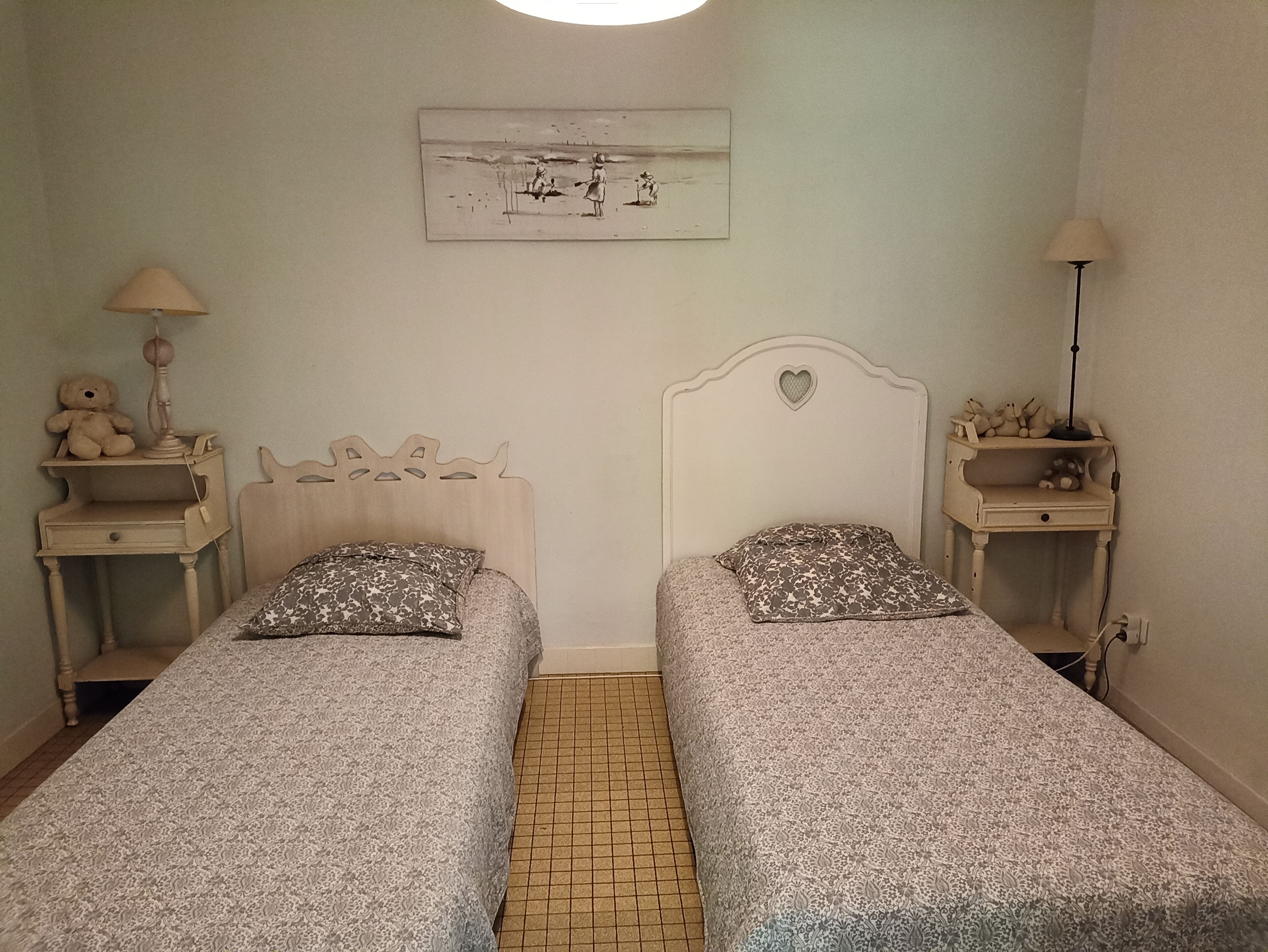 Una habitación con dos camas individuales, cada una con una colcha estampada y almohadas a juego. En cada lado de las camas hay una mesita de noche con una lámpara. Encima de las camas, un cuadro enmarcado en la pared.