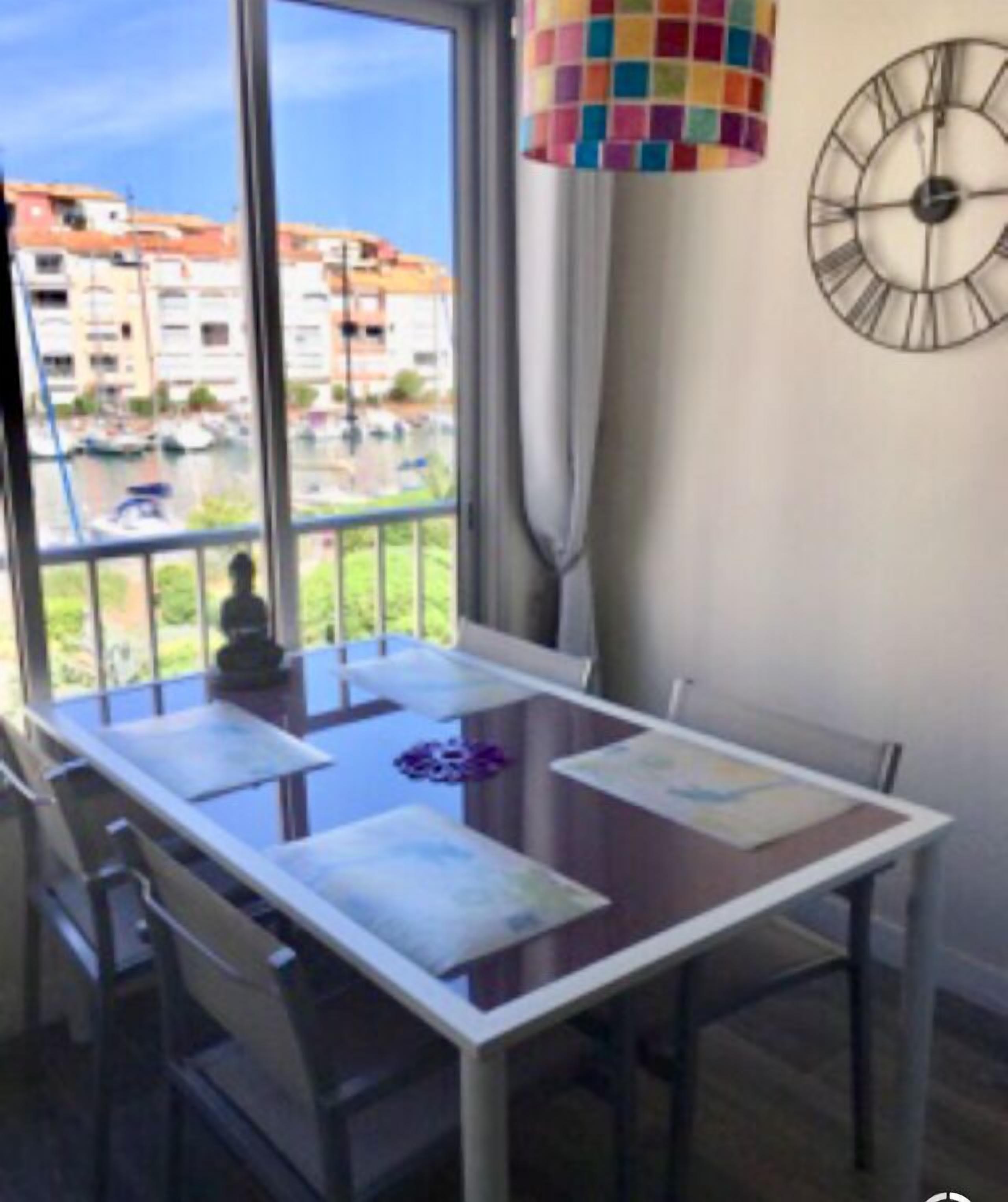 A dining area with a table and chairs, a view of buildings and a body of water outside, and a colorful hanging lamp.