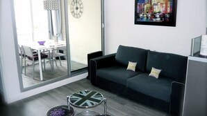 A modern living room with a grey sofa, a dining table with chairs, a glass door leading to a balcony, and a wall clock. - STUDIO RENOVATED WITH VIEW ON BOATS FOR 4 PEOPLE (Agde)