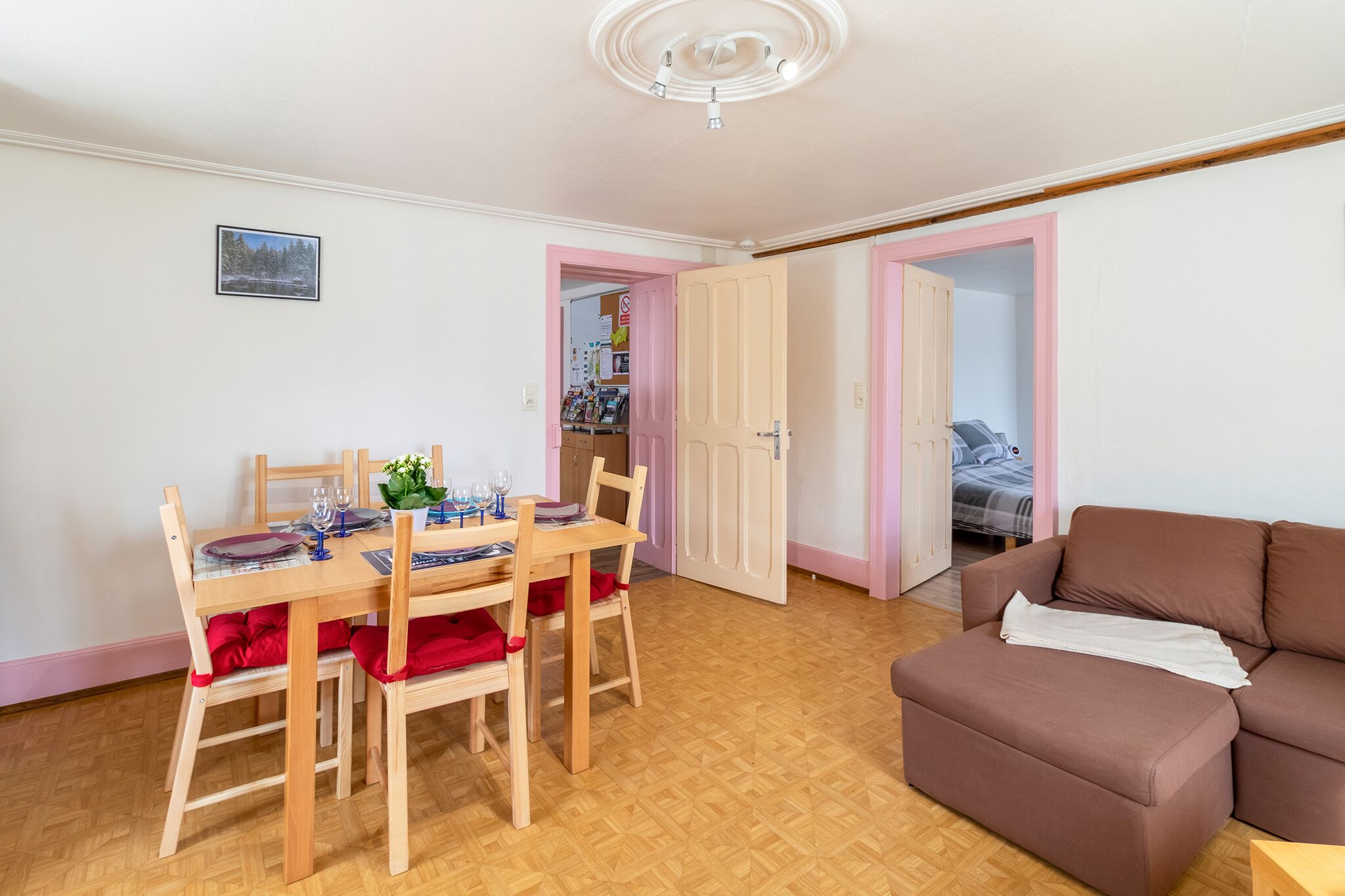 Een eetgedeelte met een houten tafel en stoelen, een bruine bank en een deur die naar een andere kamer leidt.