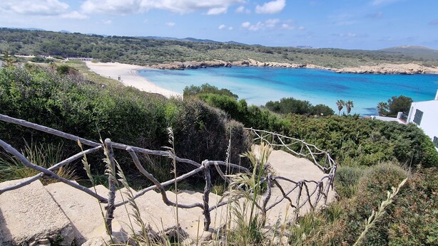 A coastal landscape with a sandy beach, clear blue waters, and a view of the sea.