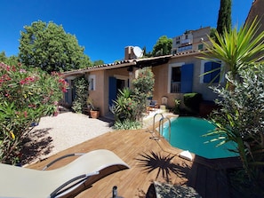Une terrasse en bois avec une piscine, entourée de plantes en pot et d’un bâtiment aux volets bleus.