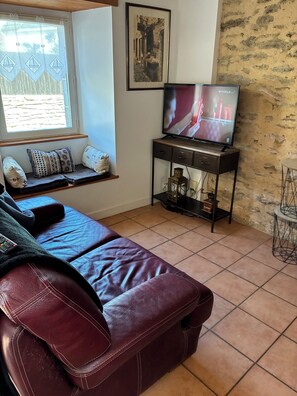 A living room with a leather sofa, a television on a stand, a window with a view, and a stone wall.