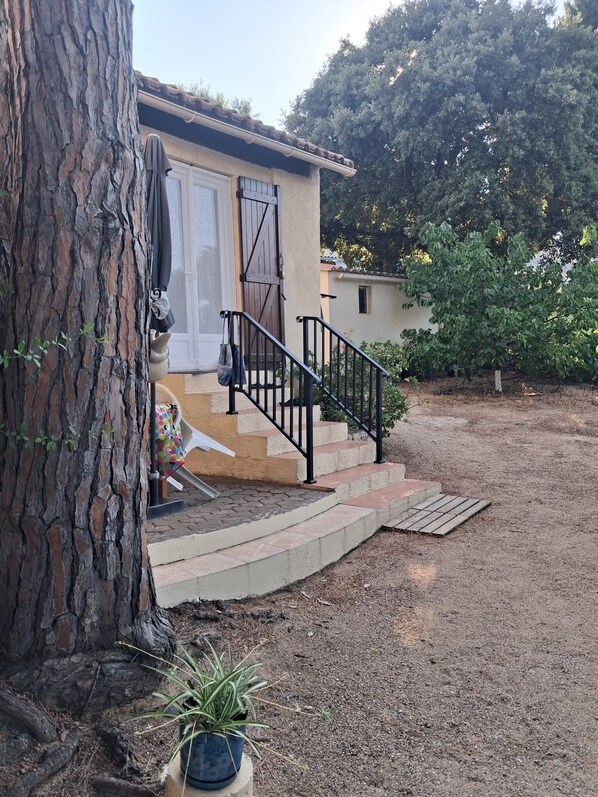 A house with a staircase leading to a door, a tree in the foreground, and a garden area. - House with garden, close to the beach, for 2 people, fully equipped. (Calvi)