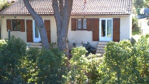 Une maison à deux étages, avec un toit en tuiles, des volets en bois et un porche à l’avant, aménagé avec des sièges d’extérieur.