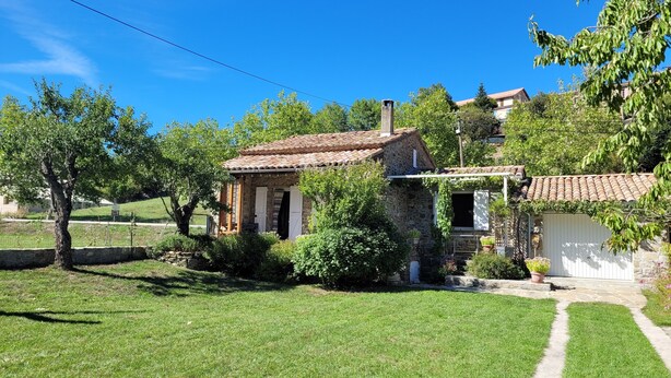 Une maison en pierre avec un toit en tuiles, entourée d’une pelouse bien entretenue et d’arbres.