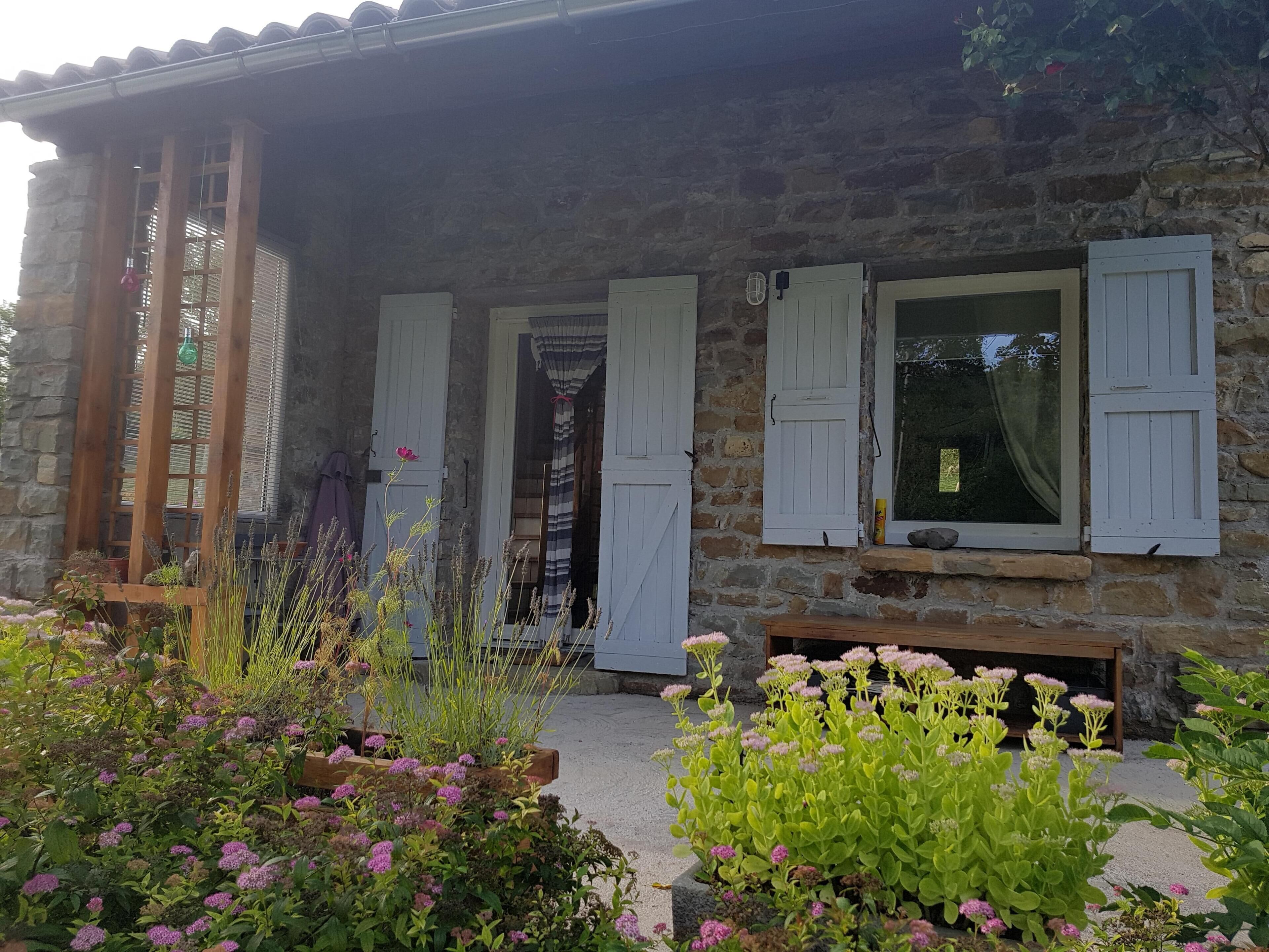 Un bâtiment en pierre avec des volets blancs, un porche en bois et un jardin où poussent différentes plantes et fleurs.