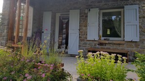 Un bâtiment en pierre avec des volets blancs, un porche en bois et un jardin où poussent différentes plantes et fleurs.