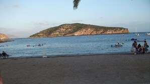Una playa con gente nadando y tomando el sol, una terraza de madera y barcos en el agua.