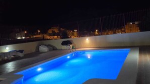 A swimming pool with a blue-lit edge, surrounded by a tiled deck and a metal railing.