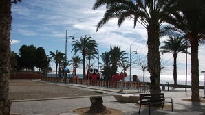 A paved walkway with benches, palm trees, and a large red sculpture.