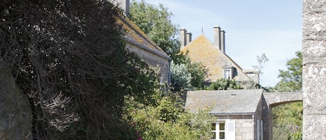 A stone archway leading to a narrow path flanked by dense vegetation and a small building with a sloped roof.