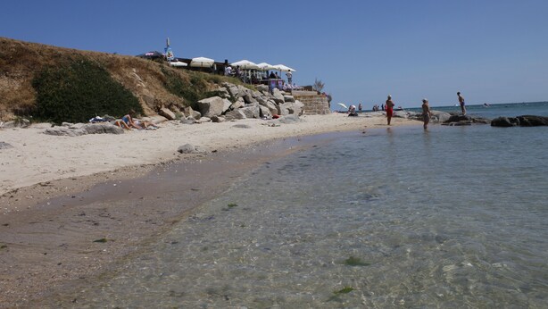 A beach with clear shallow waters, rocky outcrops, and a few people.