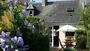 A traditional stone building with a sloped roof, a small porch with a table and chairs, and a garden with purple flowers.