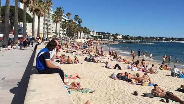 Ein belebter Strand mit badenden und sonnenden Menschen, Palmen und einem strahlend blauen Himmel.