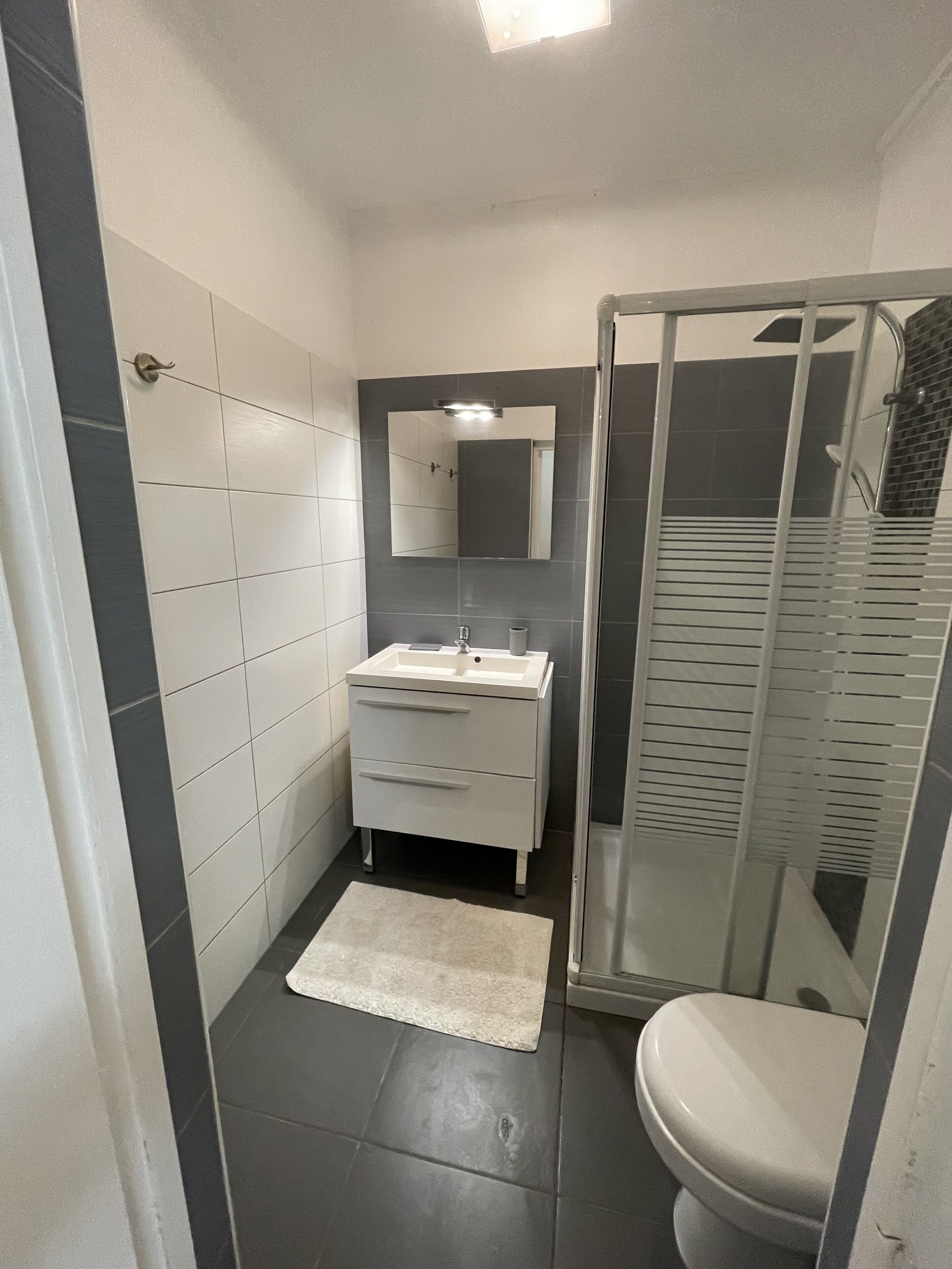 A bathroom with a white sink, mirror, and toilet, a shower with a glass door, and dark gray tiles.