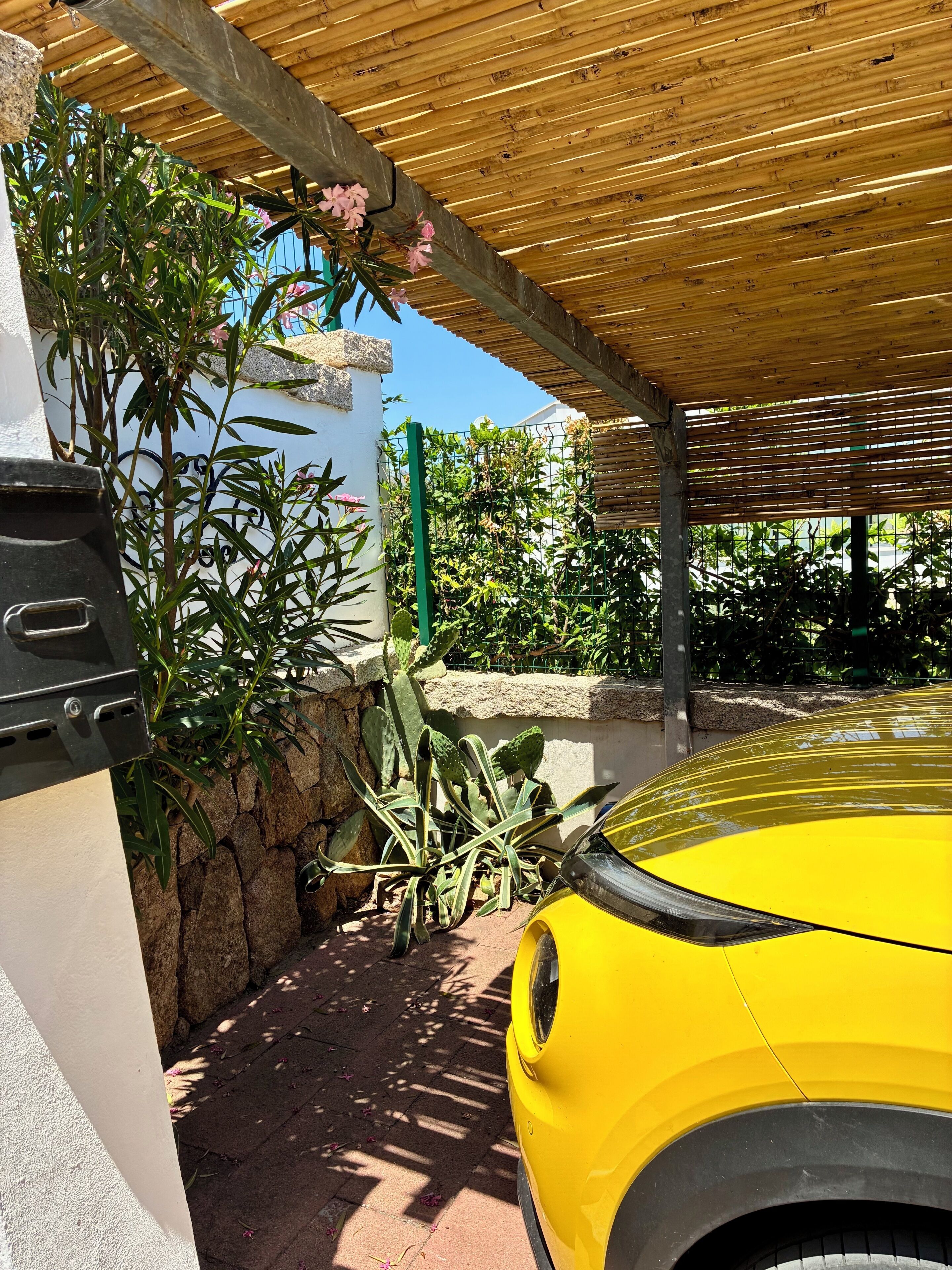 A yellow car parked under a wooden pergola with a stone wall and green plants.