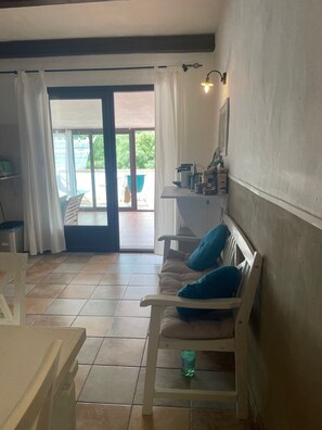 Una habitación con sofá, una mesa, una silla, un estante pequeño, una lámpara de pared y un ventanal corredero que da a un espacio exterior.