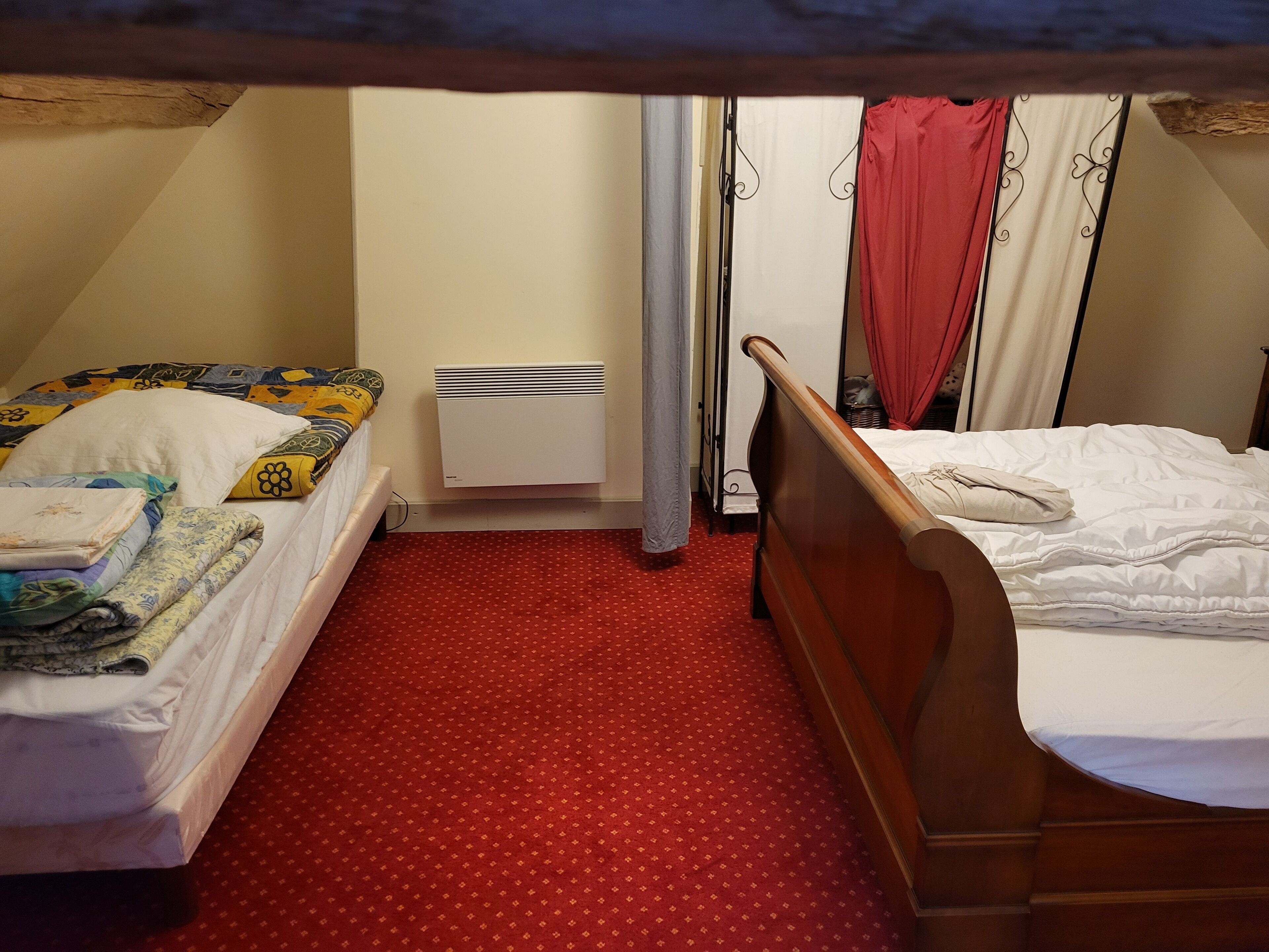 A small, cozy room with two single beds, a red patterned carpet, a white radiator, and a window with curtains.