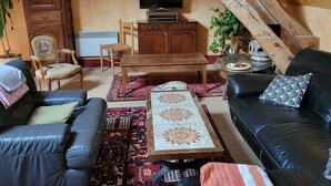 Una sala de estar acogedora con techo alto, vigas de madera, un televisor sobre un mueble de madera, un tapete estampado, un sofá de cuero negro, un armario de madera y una silla.