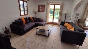 Una sala de estar con un sofá modular, una mesa de centro y una silla. Hay una ventana con cortinas, una lámpara y un cuadro en la pared.