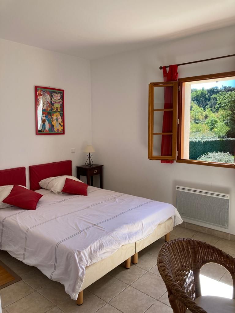 Un dormitorio con una cama grande, una silla de mimbre, una ventana con vista a follaje y un cuadro en la pared.