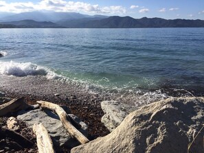 A rocky shoreline with waves crashing onto the beach, a clear sky, and mountains in the distance. - St Florent, beautiful between sea and mountain, terrace , garden, swimming pool (Saint-Florent)