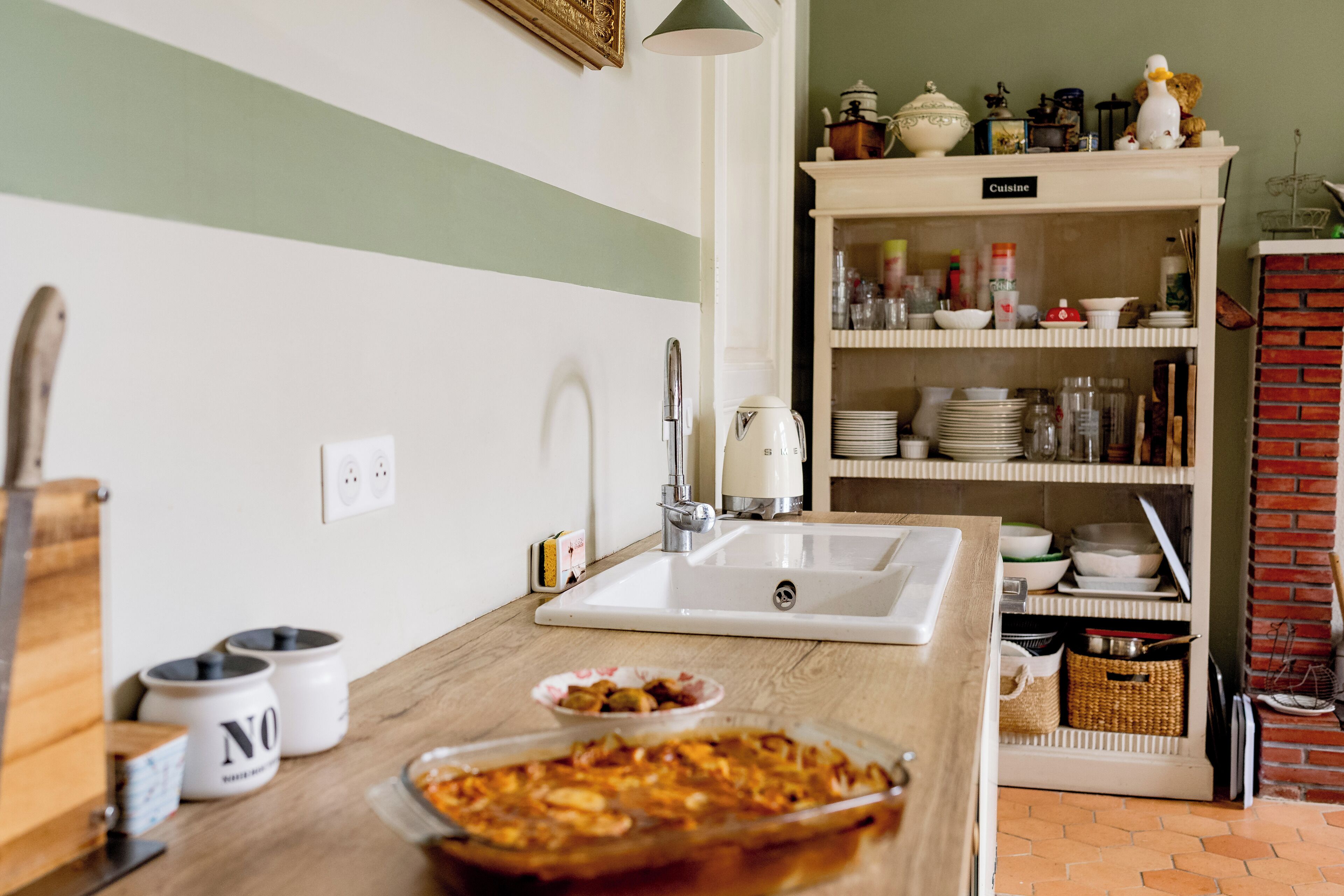 A kitchen with a white sink, wooden countertop, and a dish rack. There are jars, a kettle, and a cabinet labeled "Cuisine" in the background.