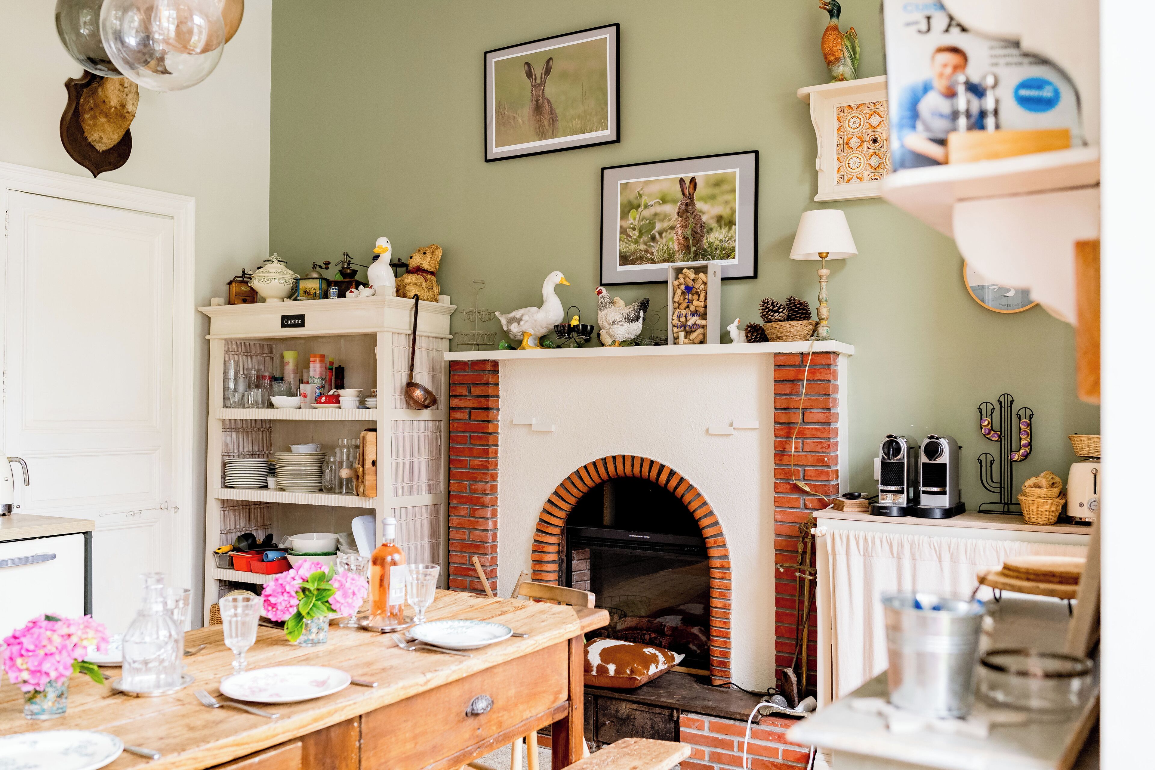 A kitchen with a brick fireplace, a dining table set with plates and glasses, and a shelf with kitchenware.