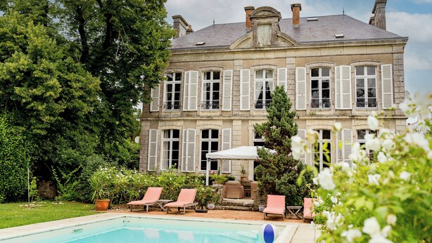 A large, historic building with a swimming pool, surrounded by lush greenery and outdoor seating.