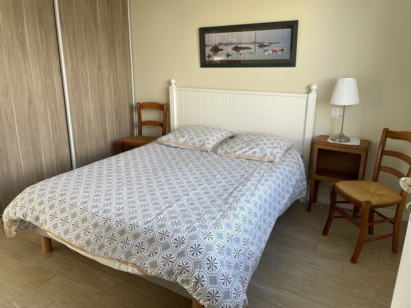 A neatly made bed with a patterned bedspread, two wooden chairs, a nightstand with a lamp, and a framed picture on the wall.
