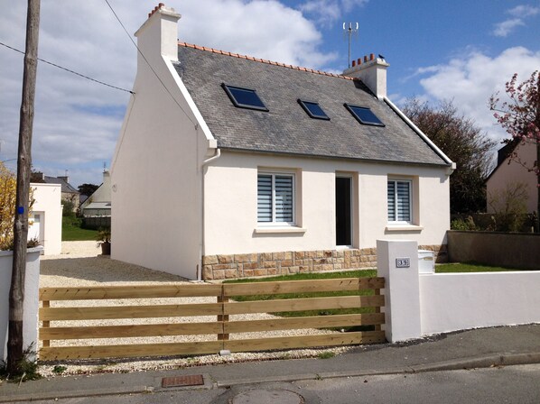 A small white house with a grey roof, two windows, and a wooden gate.
