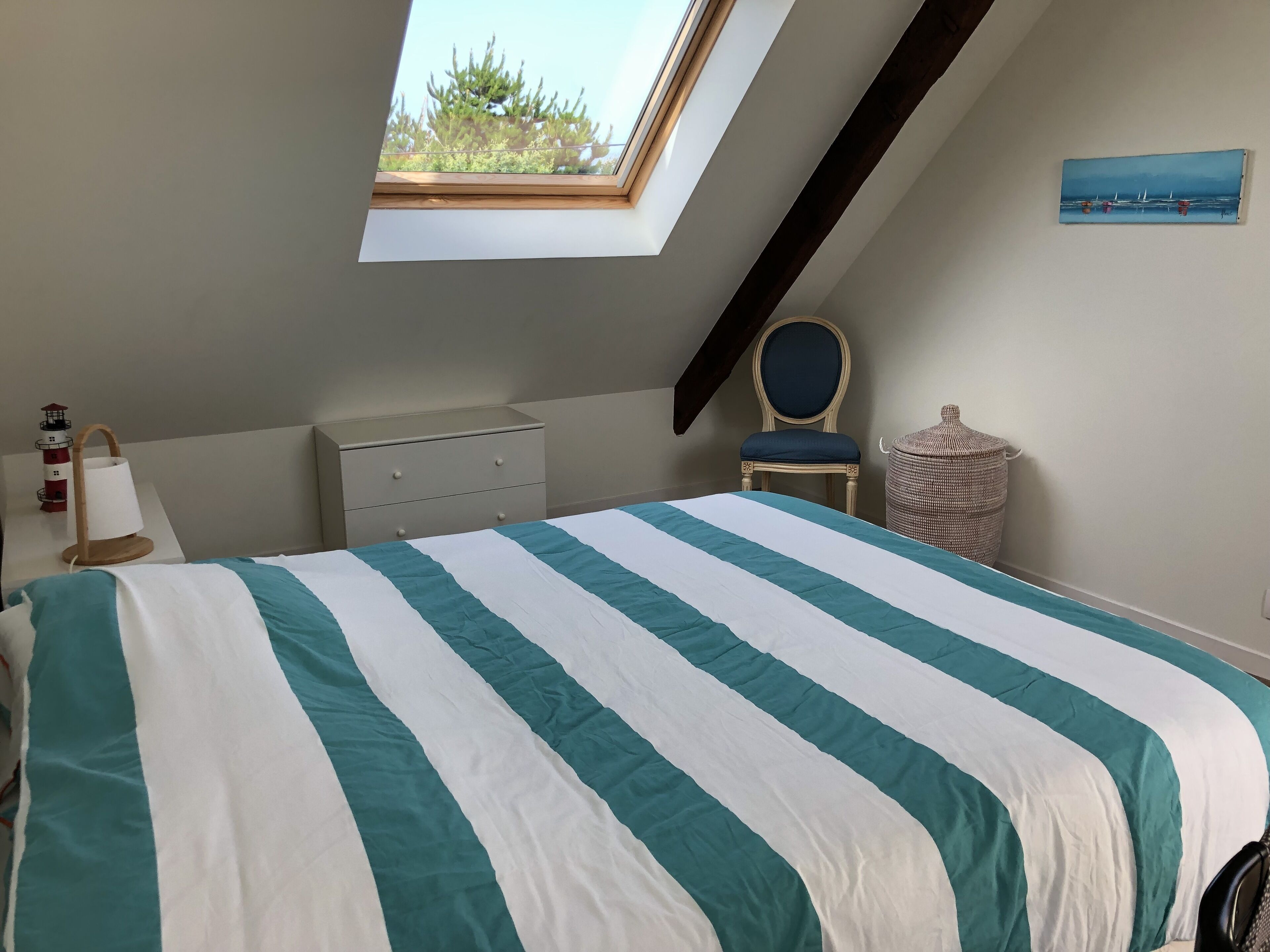 Une chambre avec un puits de lumière, un lit avec une literie à rayures, une commode, une chaise et une lampe fixée au mur.