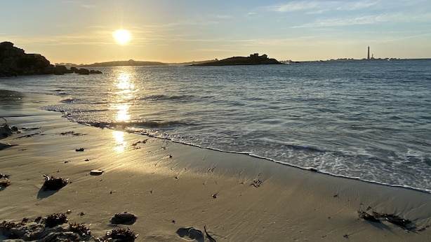 Una playa con un faro a lo lejos, agua tranquila y cielo despejado.