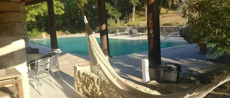A hammock under a wooden pergola overlooking a swimming pool with lounge chairs and a stone wall in the background.